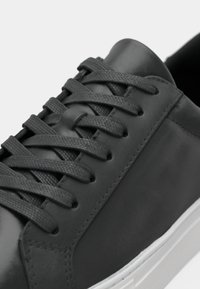Black leather sneaker with flat black laces, textured eyelets, and a white rubber sole. Smooth upper and minimalist design.