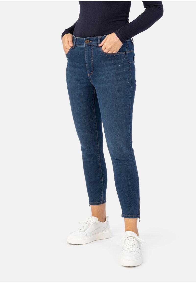 Dunkelblaue Denim-Jeans im Slim-Fit mit ausgefranstem Saum und Nietenakzenten an den vorderen Taschen. Kombiniert mit weißen Sneakers.