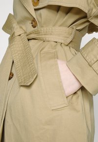 Trench-coat beige avec des boutons, ceinture nouée à la taille, et main placée dans une poche avant.