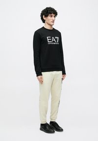 Férfi áll fekete pulóverben, fehér "EA7 Emporio Armani" logóval, krémszínű tréningnadrágban oldalsó felirattal és fekete sportcipőben, egyszínű háttér előtt.