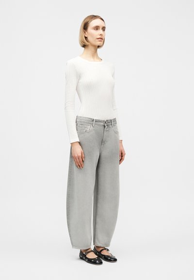 Γυναίκα που στέκεται φορώντας λευκό μακρυμάνικο ριμπ τοπ, άνετο ανοιχτό γκρι τζιν και μαύρα φλατ παπούτσια με ασημένιες λεπτομέρειες με τρύπες, μπροστά σε απλό ανοιχτό φόντο.