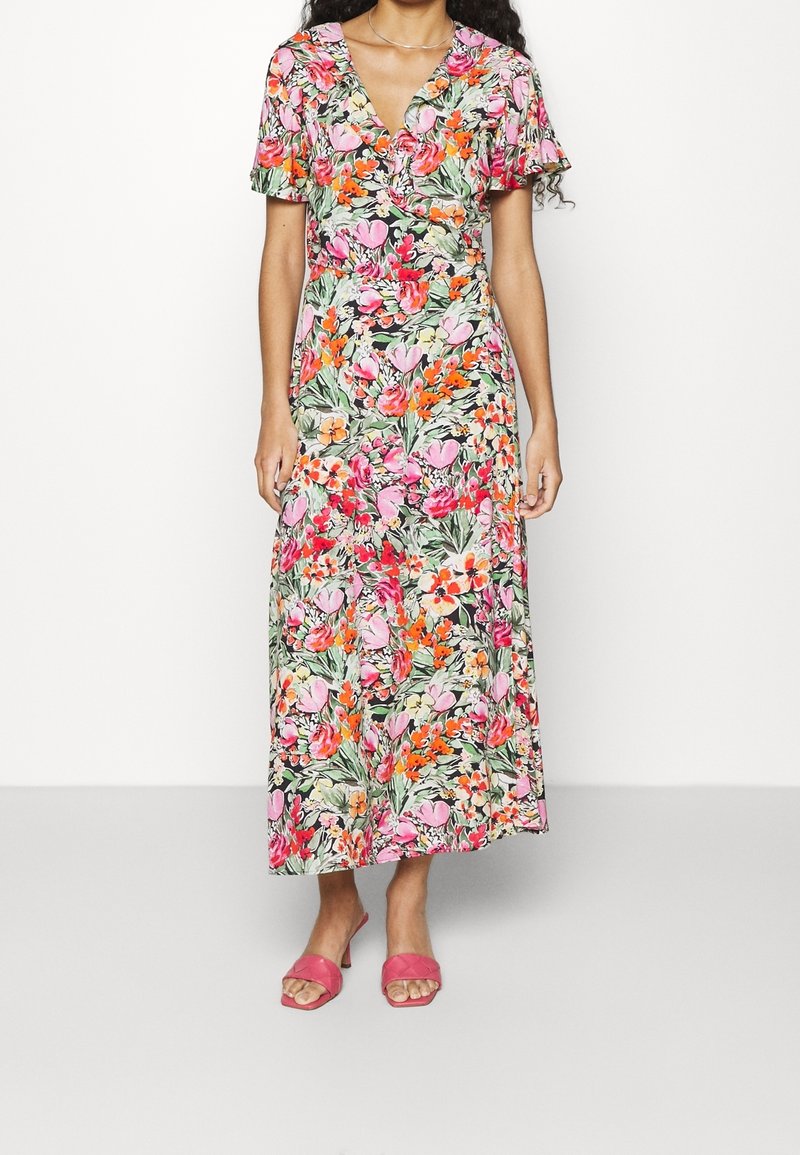 Femme portant une longue robe portefeuille florale à manches courtes et des sandales à talons roses, se tenant devant un fond clair.