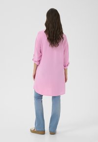 Lång rosa skjorta med upprullade ärmar, gjord av lättviktsmaterial, tillsammans med ljusblå utsvängda jeans och beige skor. Enkel och ren design.