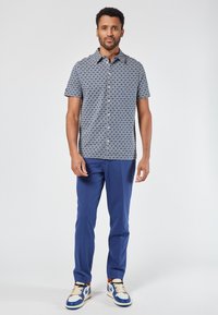 Camicia a maniche corte con un motivo geometrico blu e bianco, abbinata a pantaloni blu e sneaker multicolori. Vestibilità e design casual.