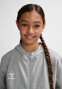 Grauer Zip-Hoodie mit weicher Textur und Kapuze. Besitzt ein kleines weißes Logo auf der linken Brust. Das Model hat lange geflochtene Haare.