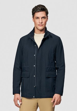 Mężczyzna w granatowej kurtce z przednimi kieszeniami i beżowych spodniach, stojący na tle jednolitego, jasnoszarego tła.