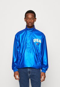 Blå, skinnende, lett vindjakke med høy krage, frontlomme og logoen "TOMMY USA" i gul og hvit. Elastiske mansjetter og nederkant.