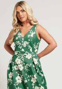 Vihreä kukkamekko, jossa on V-mallinen kaula-aukko ja leveät hihat. Siinä on valkeat kukkakuviot ja laskokset vyötäröllä, valmistettu sileästä kankaasta.