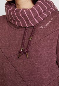 Sudadera marrón con un cuello tipo cowl a rayas, que presenta un diseño geométrico, cordones con herrajes dorados y detalles bordados.