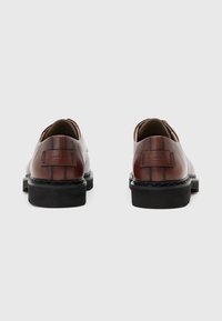 Un paio di scarpe stringate in pelle marrone con suole nere, viste da dietro su uno sfondo grigio chiaro uniforme.