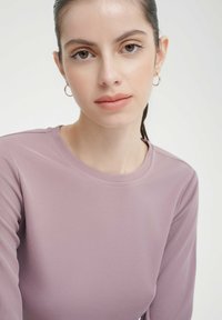Langärmliges Oberteil in hellem Mauve mit gerippter Textur. Besitzt einen runden Halsausschnitt und dezente Schulternähte und wird mit einem schlichten goldenen Creolenohrring getragen.