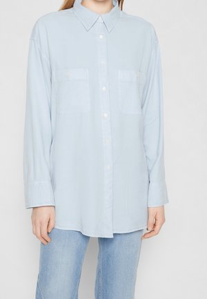 Lyseblå langærmet skjorte med knapper og to brystlommer, båret sammen med lyseblå jeans på en person mod en ensfarvet baggrund.