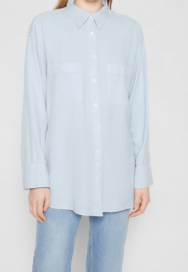 Chemise bleu clair à manches longues avec boutons et deux poches poitrine portée avec un jean bleu clair sur une personne devant un fond uni.