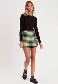 Top noir à manches courtes associé à une mini-jupe vichy verte et noire avec des boutons. Le mannequin porte des chaussures noires épaisses avec des accents argentés.