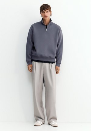 Un jeune homme se tient contre un mur blanc, portant un sweat gris à fermeture éclair quart, un pantalon ample beige clair et des baskets blanches, face à l'avant.