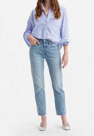 Femme portant une chemise à rayures bleu clair boutonnée, un jean taille haute bleu clair et des ballerines argentées, debout avec une main dans la poche.
