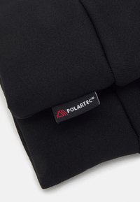 Tissu Polartec noir avec une texture lisse, présentant une étiquette avec logo visible et des accents rouges sur le bord. Design plié mettant en valeur le matériau en couches.
