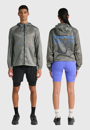 Zwei Personen tragen graue Windjacken mit Kapuze; eine schaut nach vorne und trägt schwarze Shorts, die andere schaut nach hinten und trägt lila Radsport-Shorts und weiße Socken.