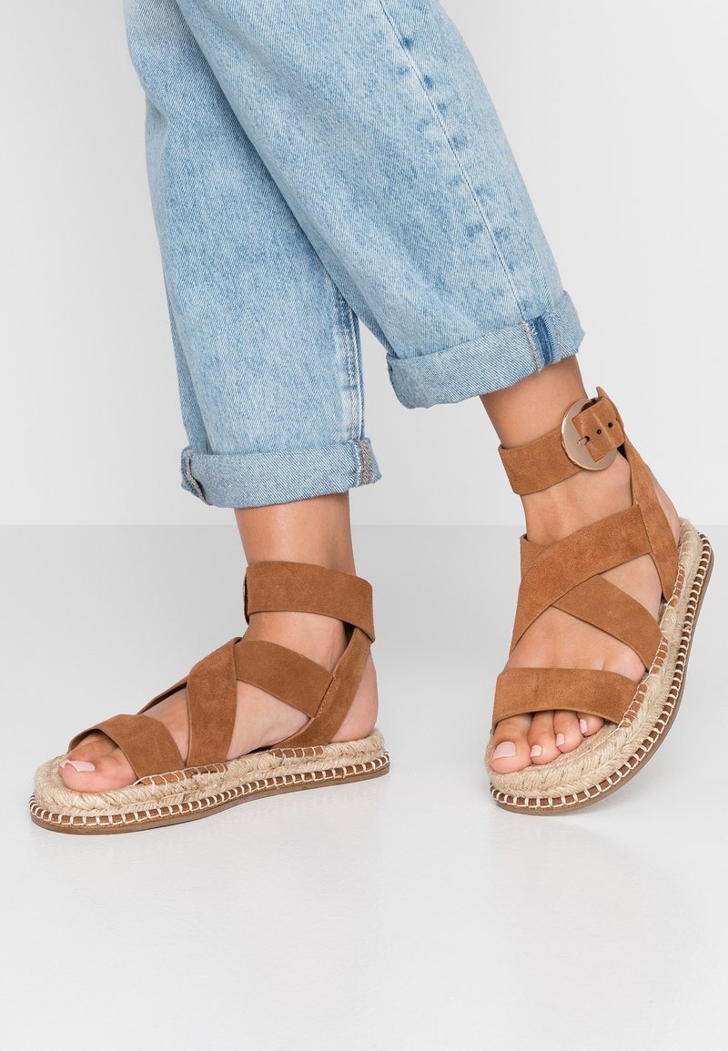 Bruna mockaskinns sandaler med remmar och en jutesula, med ett brett ankelsnöre och spänne, parat med uppvikta ljusblå jeans.