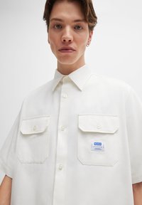 Weißes Kurzarmhemd mit Button-Down-Kragen, zwei Vorderaschen und einem blauen "HUGO"-Label an der linken Tasche. Glatte Stofftextur.