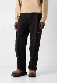 Black cargo pants with two side pockets, textured fabric, and a loose fit. Worn with brown suede slip-on shoes. Beige top partially visible.