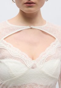 Λευκό τοπ από δαντέλα με λουλουδένια σχέδια, βαθιά V λαιμόκοψη με διακοσμητικό κλείσιμο κουμπιών και διαφανή μακριά μανίκια για πολυεπίπεδη υφή.