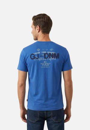 Uomo che indossa una maglietta blu a maniche corte con testo stampato sulla schiena e jeans blu scuro, in piedi di spalle.