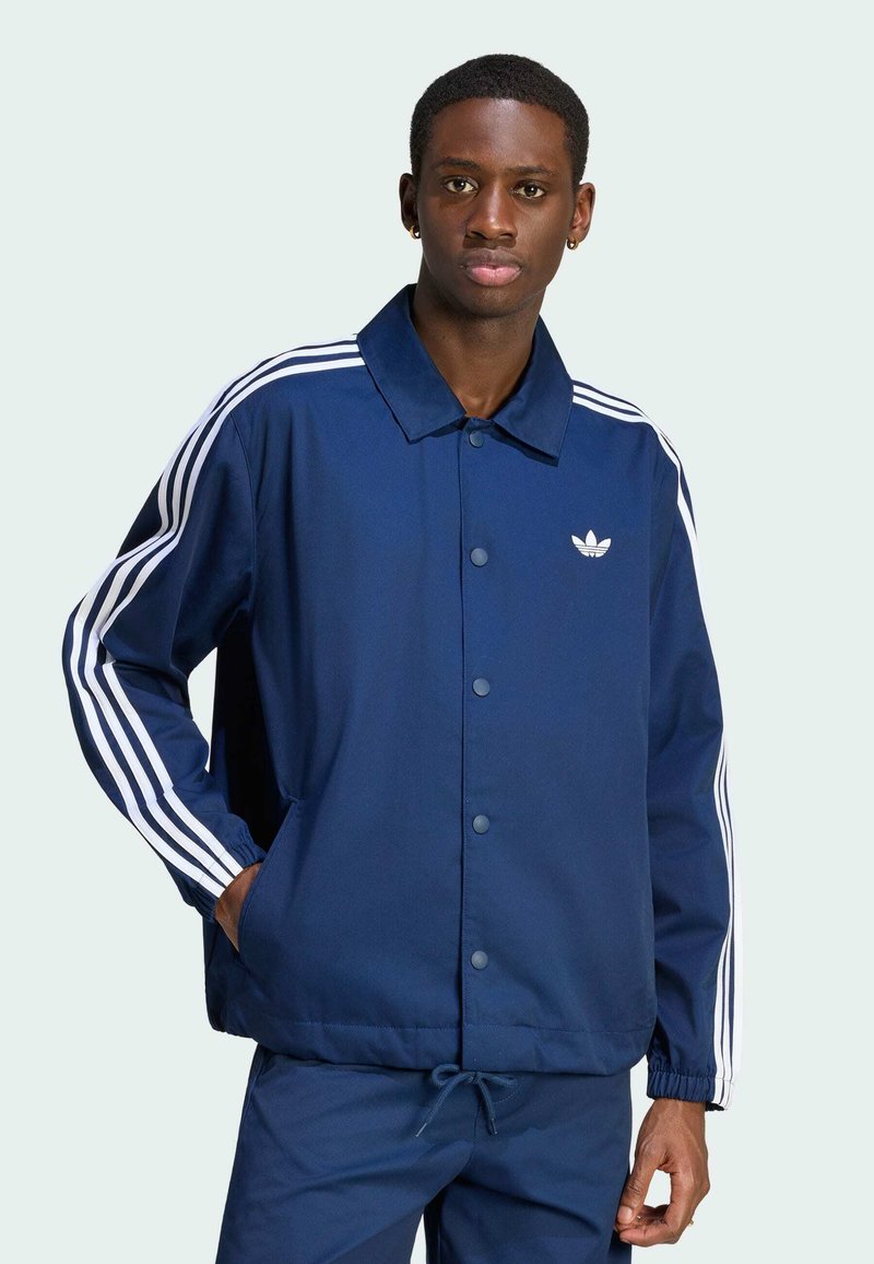 Jeune homme portant une veste Adidas bleue avec des bandes blanches sur les manches et les poches, debout avec une main dans la poche devant un fond uni.