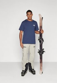 Navy majica s logotipom, svijetlosive hlače s crnim detaljima i crne skijaške čizme; osoba drži smeđe skije s vezovima.