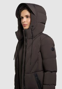 Brun puffjacka med stor huva, framdragkedja, sidofickor och en logotyp på vänster ärm. Strukturerad tyg med en quiltad design.