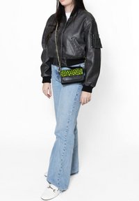 Giacca bomber nera in pelle, jeans svasati azzurri chiari e una borsa a tracolla nera con un accento a stampa leopardata verde brillante. Sneakers bianche.