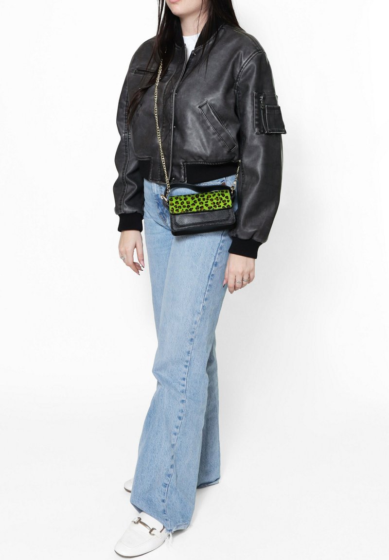 Giacca bomber nera in pelle, jeans svasati azzurri chiari e una borsa a tracolla nera con un accento a stampa leopardata verde brillante. Sneakers bianche.
