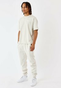 Camiseta de manga corta beige y pantalones deportivos a juego, ambos con el texto blanco "PROJECT X PARIS". Estilizados con zapatillas blancas.