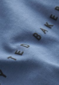 Svetlo modra tkanina z dvignjenimi črnimi črkami, ki črkujejo "TED BAKER," poudarja gladko teksturo in subtilen lesk.