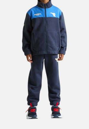 Bambino che indossa una tuta blu navy e blu con cerniera, polsini elastici e scarpe da ginnastica nere con lacci rossi su sfondo bianco.