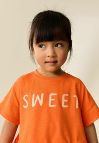 Narancssárga rövid ujjú póló, világos rózsaszín "SWEET" felirattal. Puha, texturált anyag.