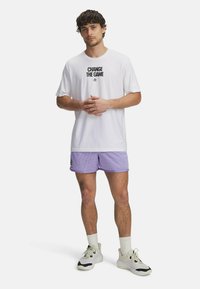 Camiseta blanca de manga corta con "CAMBIA EL JUEGO" impreso en negro. Pantalones cortos de malla lavanda. Tejido ligero, corte relajado, combinados con zapatillas blancas.