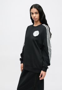 Μαύρο φούτερ με λευκές ρίγες κατά μήκος των μανικιών, με κυκλικό λογότυπο σε ασημί, κατασκευασμένο από βαμβάκι με απαλή υφή.