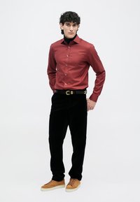 Muškarac s kovrčavom kosom nosi bordo košulju s dugim rukavima preko crnog dukserice, crne hlače, crni remen i smeđe tenisice, stoji nasuprot bijeloj pozadini.