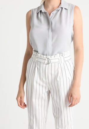 Femme portant un chemisier sans manches gris clair et un pantalon blanc taille haute à fines rayures verticales noires avec une ceinture assortie.