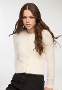Cardigan bianco e soffice con scollo rotondo e maniche lunghe, caratterizzato da bottoni decorativi lungo la parte anteriore. Progettato per un look aderente.