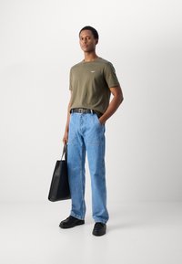 Oliven grøn bomulds t-shirt med et lille logo, blå denim jeans med en afslappet pasform, sort bælte, sorte sko og en sort totebag.