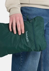 Grön vadderad påse med en jämn yta, försedd med dragkedjestängning och ett märkeslogotyp. Hand som synligt håller i påsen.