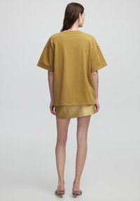 T-shirt surdimensionné jaune moutarde à manches courtes, avec un ourlet droit, porté sur une courte jupe dorée satinée. Texture lisse, coupe décontractée.