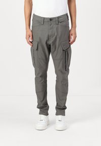 Γκρίζες cargo παντελόνες με στενή εφαρμογή, εφοδιασμένες με αρκετές τσέπες, ενισχυμένες ραφές και ρυθμιζόμενους μανικέτες, συνδυασμένες με λευκά αθλητικά παπούτσια.