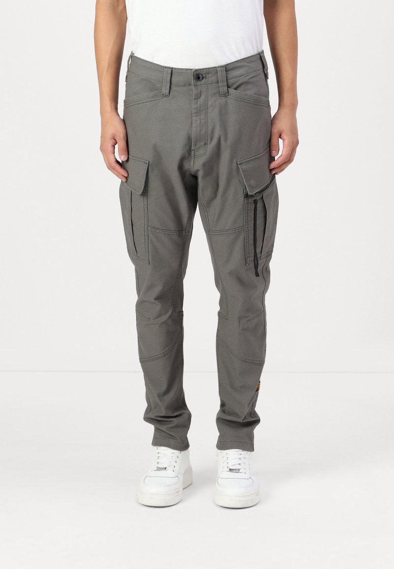 Γκρίζες cargo παντελόνες με στενή εφαρμογή, εφοδιασμένες με αρκετές τσέπες, ενισχυμένες ραφές και ρυθμιζόμενους μανικέτες, συνδυασμένες με λευκά αθλητικά παπούτσια.