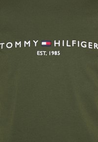 Grünes Baumwoll-T-Shirt mit dem Schriftzug "TOMMY HILFIGER" in weißer Stickerei und einer Flaggengrafik, sowie "EST. 1985" darunter in weißem Text.
