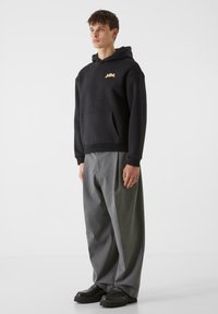 Črn bombažna kapuca s sprednjim žepom in zlatim logotipom, v kombinaciji s sivimi širokimi hlačami in črnimi robustnimi čevlji. Enostaven in sodoben dizajn.