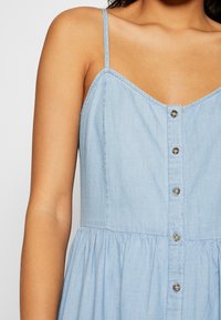 Ljusblå chambraydress med V-ringning, tunna axelband och knappslå framtill. Har en rynkad midja för en avslappnad passform och mjuk textur.