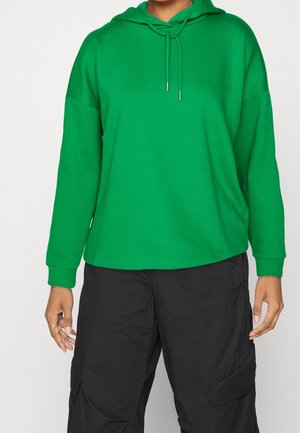 Person wearing a bright green hoodie with drawstrings and loose black cargo pants, standing against a plain background.
